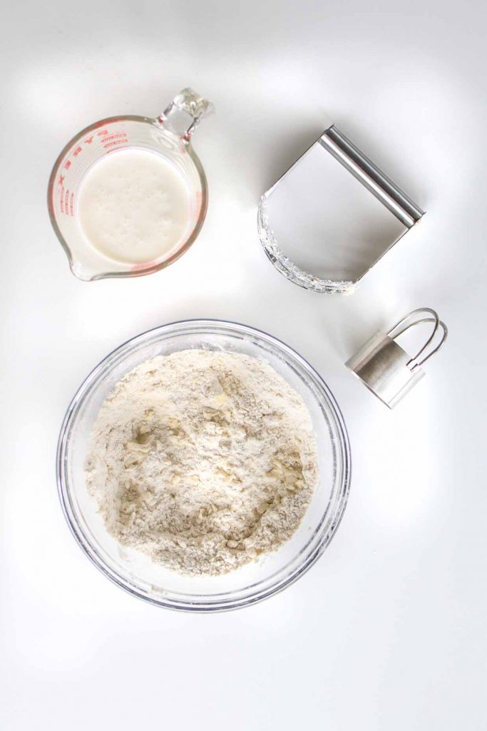 aerial view of bowl of flour, cup of buttermilk, pastry cutter and biscuit cutter
